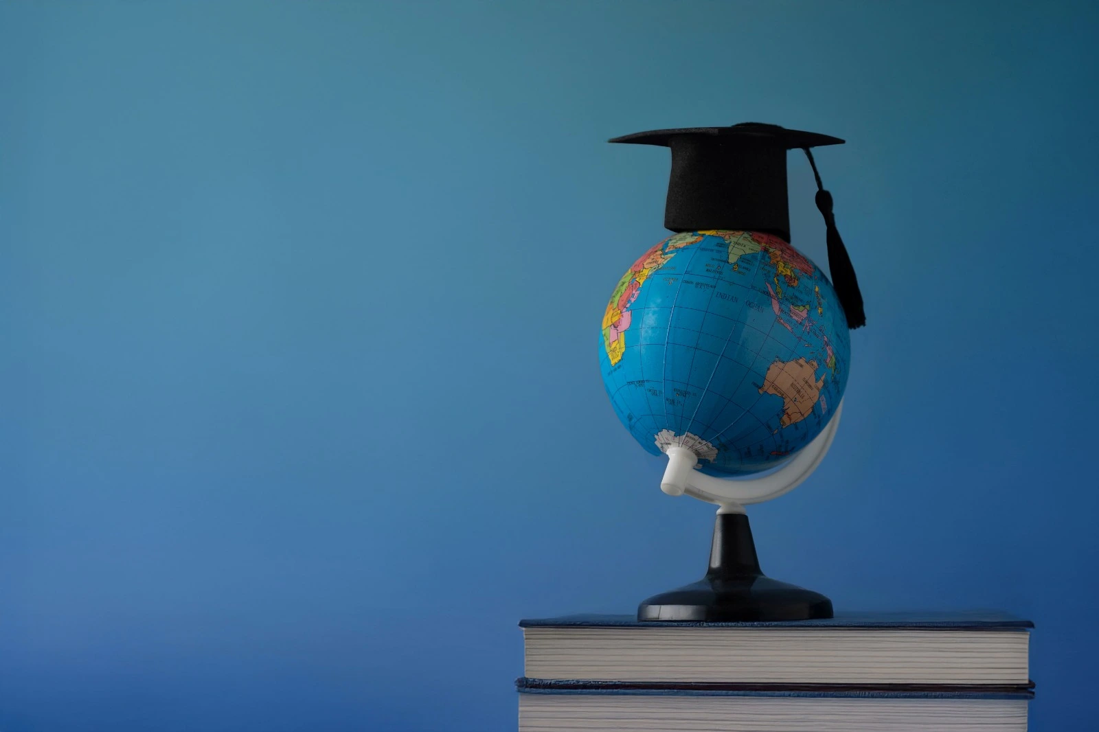 Small world globe wearing a black graduation cap sitting on top of books against a gradient blue background.