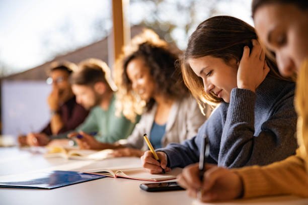 Side view close-up of a young woman in a gray sweater writing in a notebook with a pen, sitting in a row of blurred students doing the same.